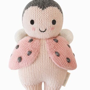 May include: A knitted ladybug plush toy in soft pink, cream, and gray. The toy has a smiling face, small antennae, and wings with gray spots. The plush is designed for children.