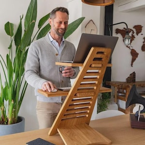 Puede incluir: Un escritorio de pie de madera con un portátil, un ratón y una taza de café. El escritorio tiene un diseño de listones y está hecho de madera de color claro. Una persona está de pie junto al escritorio. Un cuaderno y un bolígrafo están sobre la mesa.