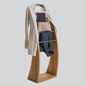 May include: A wooden clothing rack with a curved design, holding a beige button-down shirt, navy blue pants, tan pants, and dark blue pants. The rack has three metal bars for hanging clothes.