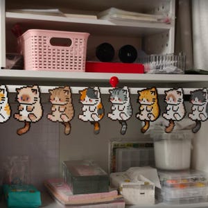 May include: A collection of nine pixelated cat ornaments in various colors, including orange, brown, gray, and white, are hanging from a black bar. The cats are designed with a sitting pose and have long tails. The background shows a white shelf with storage items.