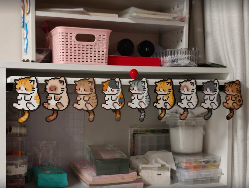 May include: A collection of nine pixelated cat ornaments in various colors, including orange, brown, gray, and white, are hanging from a black bar. The cats are designed with a sitting pose and have long tails. The background shows a white shelf with storage items.