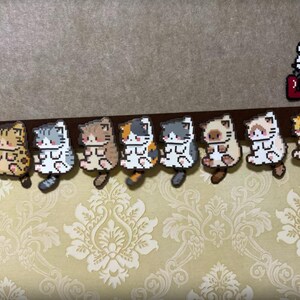 May include: A collection of pixelated cat figurines in various colors, including gray, brown, and orange, arranged in a row. A Hello Kitty design with the word "LOVE" is also present. The figurines are attached to a brown surface.