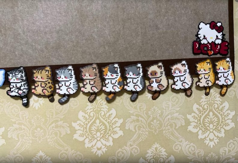 May include: A collection of pixelated cat figurines in various colors, including gray, brown, and orange, arranged in a row. A Hello Kitty design with the word "LOVE" is also present. The figurines are attached to a brown surface.