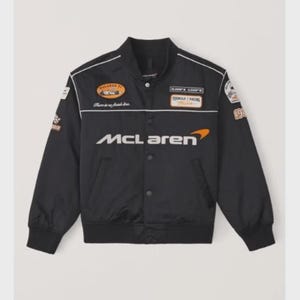 Può includere: Giacca da corsa McLaren nera con dettagli bianchi e varie toppe. Presenta il logo McLaren in bianco e arancione. Include il testo "Formula 1 Racing" e "McLaren F1".