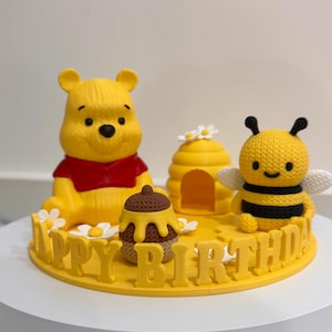 以下が含まれることがあります： 赤いシャツを着た黄色いクマのフィギュア、黄色い蜂の巣、茶色の蜂蜜の壺、黒い縞模様と白い羽を持つ黄色いハチのフィギュア。これらはHAPPY BIRTHDAYという文字が刻まれた黄色い円形の台座の上に配置されています。