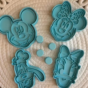 Può includere: Una collezione di formine per biscotti azzurre raffiguranti i volti di Topolino, Minnie, Pippo e Paperina. Sono inclusi anche diversi piccoli oggetti rotondi azzurri. Gli oggetti sono disposti su una superficie bianca tessuta.