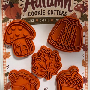 Può includere: Un set di cinque tagliabiscotti in plastica arancione su un cartoncino con la scritta Autumn Cookie Cutters. Le forme includono una casa a fungo, un maglione lavorato a maglia, una foglia d'acero, una tazza e una ghianda. Testo Made with love.