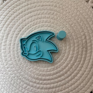 Può includere: Un tagliabiscotti azzurro a forma di Sonic the Hedgehog, con un piccolo cilindro blu abbinato. Il tagliabiscotti presenta i capelli a spillo e il volto sorridente del personaggio. Gli oggetti sono su una superficie strutturata bianco sporco.