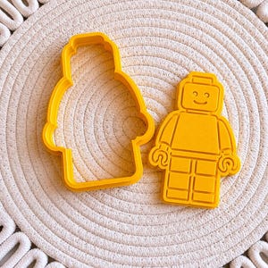 Può includere: Due formine per biscotti in plastica gialla a forma di personaggio Lego. Una è il contorno, l'altra una versione dettagliata. Sono su un tappetino bianco intrecciato.