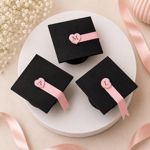 Può includere: Tre tochi di laurea neri quadrati con nastri rosa e dettagli a forma di cuore. Ogni cuore presenta una lettera: M, A o L. I tochi hanno una finitura testurizzata e sono disposti su una superficie bianca circolare con nastri rosa.