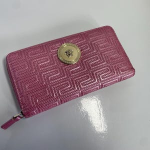 May include: A pink leather wallet with a geometric pattern and a gold-colored medallion. The wallet has a zipper closure and a rectangular shape. The medallion features a detailed design.