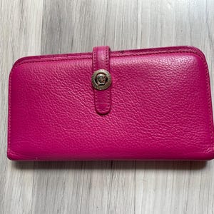 Może przedstawiać: Jasnoróżowy skórzany portfel o teksturowanej powierzchni. Portfel posiada zapięcie na klapę z okrągłym, srebrnym emblematem. Portfel jest prostokątny i wydaje się być zamknięty.