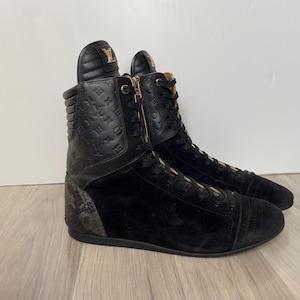 May include: Pair of black high-top trainers with gold detailing. The trainers have a quilted leather upper with embossed logos, suede panels, and a textured heel. They feature a zip closure and gold-coloured laces.