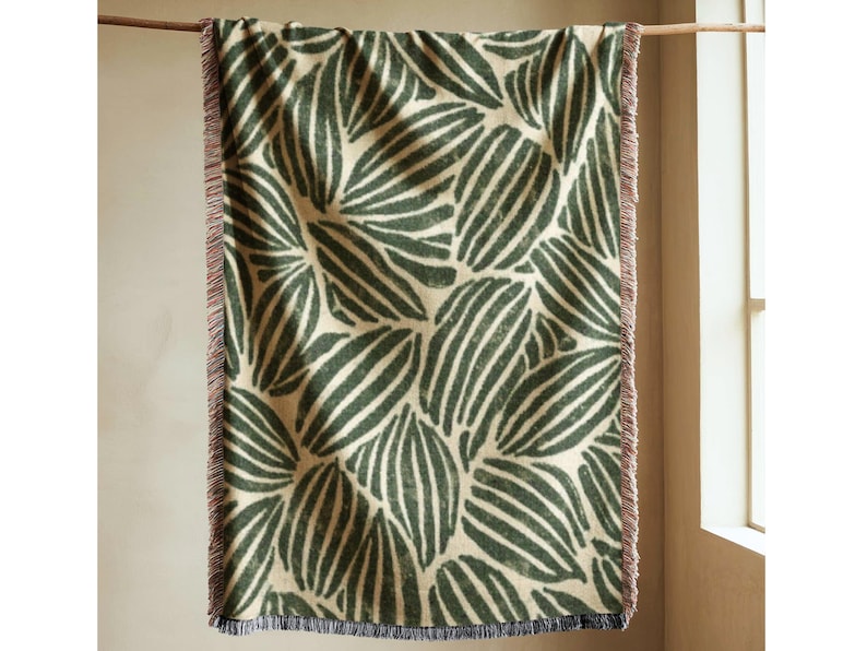 Green woven cotton blanket with repeating leaf pattern in deep forest tones on a soft neutral background, styled as a wall hanging in a warm minimal interior with natural light, textured fringe edges, and calm earthy decor, modern botanical textile throw for sofa or bed.