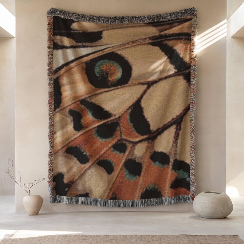 Op de afbeelding: Een rechthoekige deken met een detailpatroon van een vlindervleugel. Het ontwerp toont tinten van beige, bruin en zwart met gedetailleerde patronen. De deken heeft een gefranjerde rand. Deze decoratieve deken is ideaal om een vleugje door de natuur ge&iuml;nspireerde kunst aan elke kamer toe te voegen.