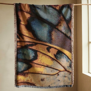 Op de afbeelding: Een rechthoekige sprei met een vlindervleugelontwerp. De sprei heeft een gedetailleerd patroon in tinten bruin, geel, blauw en paars. De randen hebben een franjes, en de sprei hangt aan een houten roede.