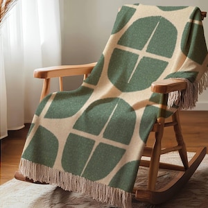 May include: A woven throw blanket featuring a repeating pattern of dark green semi-circular shapes on a cream background. The blanket has a textured appearance and is finished with long, cream-coloured fringe along the bottom and top edges.