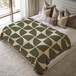 Könnte beinhalten: Eine grüne und beige geometrisch gemusterte Wurkdecke mit Fransen. Die Decke ist über einem Bett mit Kissen drapiert. Das Muster besteht aus Halbkreisen in einem sich wiederholenden Design. Die Decke ist aus einem weichen Material.