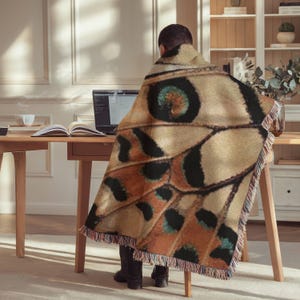 Op de afbeelding: Een geweven plaid met een gedetailleerd vlindervleugelpatroon in tinten beige, oranje en zwart met een opvallende cirkelvormige oogvlek. De rechthoekige deken heeft een gefranjerde rand en is gedrapeerd over de rug van een zittend persoon.