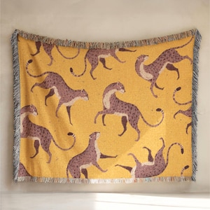 Peut inclure: Une couverture tissée avec un motif répété de guépards stylisés dans des tons de marron et de violet sur un fond jaune. La couverture a une bordure frangée grise et blanche. Cette couverture décorative convient à un lit ou un canapé.