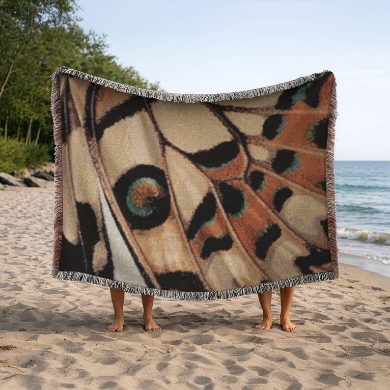 Op de afbeelding: Een geweven sprei met een vlindervleugelpatroon in tinten beige, bruin, zwart en turquoise. De deken heeft een gefranjerde rand. De afbeelding toont de deken die op een zandstrand met de zee op de achtergrond wordt vastgehouden.