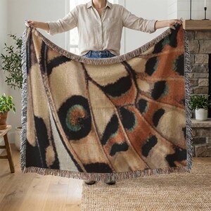 Op de afbeelding: Een grote sprei met een gedetailleerd vlindervleugelpatroon in tinten beige, bruin, zwart en teal. De sprei heeft een gefranjerde rand. De sprei wordt door een persoon omhoog gehouden.