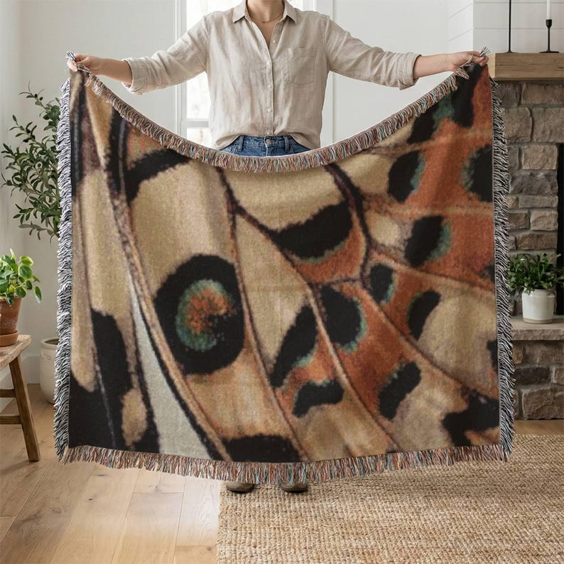 Op de afbeelding: Een grote sprei met een gedetailleerd vlindervleugelpatroon in tinten beige, bruin, zwart en teal. De sprei heeft een gefranjerde rand. De sprei wordt door een persoon omhoog gehouden.