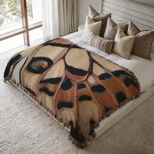Op de afbeelding: Een geweven plaid met een gedetailleerde afbeelding op grote schaal van een vlindervleugel in tinten beige, oranje, zwart en groenblauw. De rechthoekige deken heeft een rand met franjes en ligt over een bed met wit beddengoed en kussens.