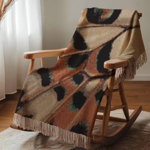 Op de afbeelding: Een patroon deken op een houten schommelstoel. De deken toont een vlindervleugelontwerp in bruin, beige, zwart en blauwgroen. De deken heeft een gefranjerde rand en is over de stoel gedrapeerd.