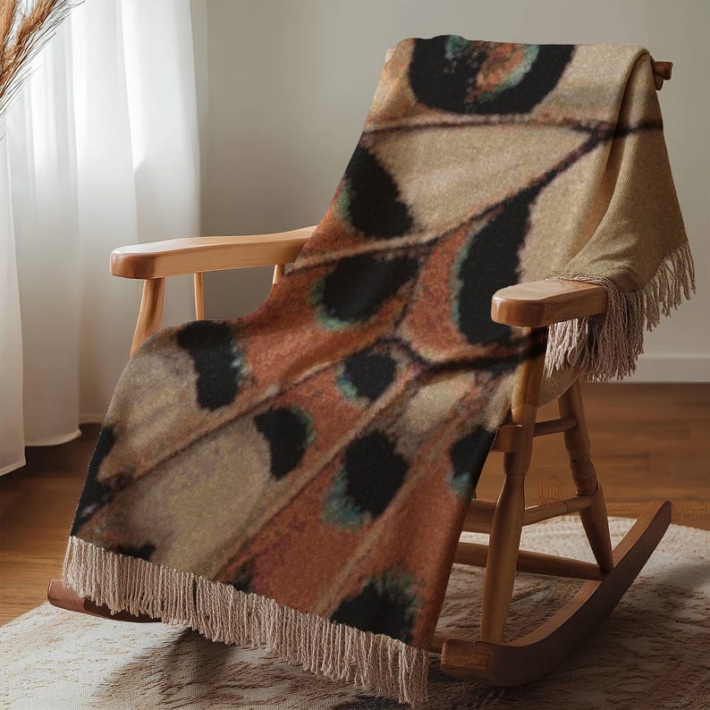 Op de afbeelding: Een patroon deken op een houten schommelstoel. De deken toont een vlindervleugelontwerp in bruin, beige, zwart en blauwgroen. De deken heeft een gefranjerde rand en is over de stoel gedrapeerd.