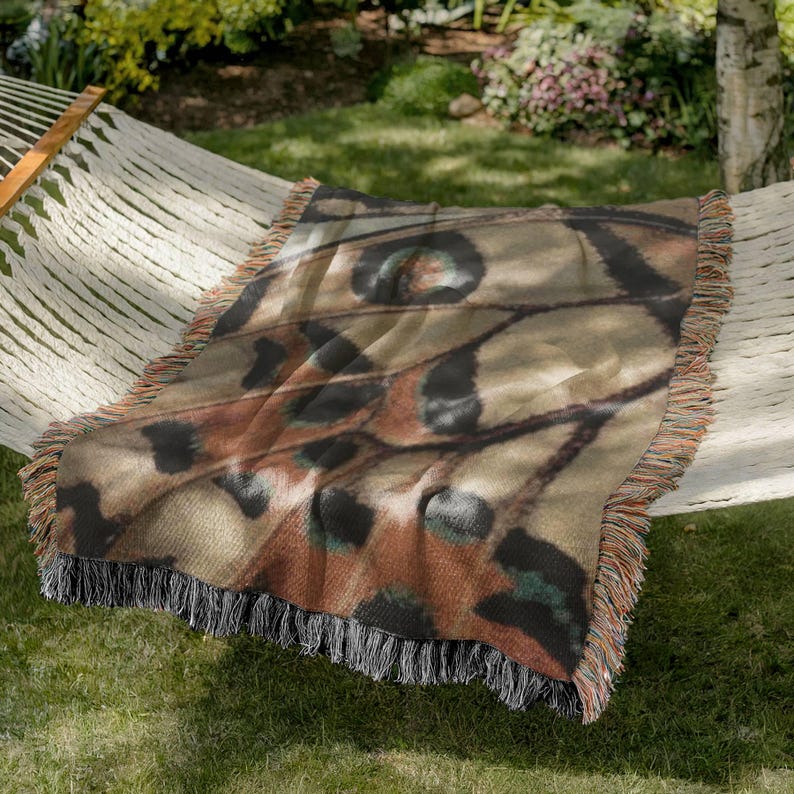 Op de afbeelding: Een rechthoekige sprei met een vlindervleugelpatroon in bruin, zwart en groen. De sprei heeft een gefranjerde rand en is over een hangmat gedrapeerd. De sprei is gemaakt van een zacht materiaal.