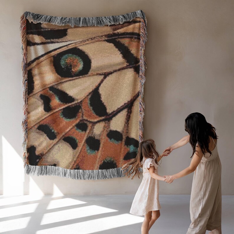 Op de afbeelding: Een geweven sprei met een vlindervleugelpatroon in tinten bruin, zwart en blauwgroen. De deken, met een gefranjerde rand, wordt aan een muur getoond. Het ontwerp is een close-up van een vlindervleugel.