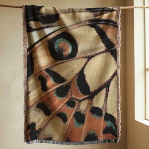 Abstract woven throw blanket featuring a butterfly wing pattern in warm neutral tones of oat, rust, charcoal and muted teal. Large scale jacquard throw blanket with symmetrical wing geometry and rich woven texture, styled hanging vertically to show full pattern and fringed edges. Statement woven blanket for modern home decor, art inspired textile design, neutral interior styling, and contemporary living room or bedroom decor.