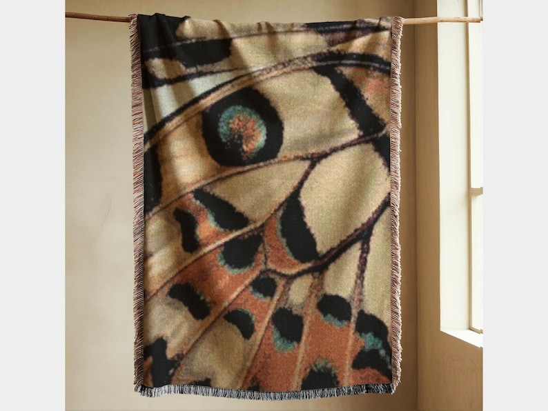 Abstract woven throw blanket featuring a butterfly wing pattern in warm neutral tones of oat, rust, charcoal and muted teal. Large scale jacquard throw blanket with symmetrical wing geometry and rich woven texture, styled hanging vertically to show full pattern and fringed edges. Statement woven blanket for modern home decor, art inspired textile design, neutral interior styling, and contemporary living room or bedroom decor.