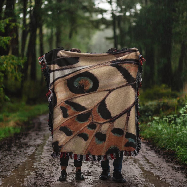 Op de afbeelding: Een beige en zwarte deken met een vlindervleugelpatroon en rode en zwarte franjes. De deken is over twee mensen gedrapeerd die op een modderig pad in een bos staan. Het ontwerp van de deken bevat bruine, zwarte en oranje details.