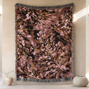 Op de afbeelding: Een geweven sprei met een botanisch patroon in tinten bruin, zwart, groen en roze. De deken heeft een gefranjerde rand en wordt tentoongesteld tegen een neutrale muur. Het ontwerp bevat abstracte bloemelementen.