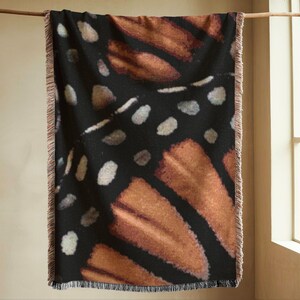 Op de afbeelding: Een rechthoekige sprei met een patroon van een monarchvlindervleugel. De deken heeft een zwarte achtergrond met oranje en bruine vleugeld details en witte stippen. De randen hebben een franjes.
