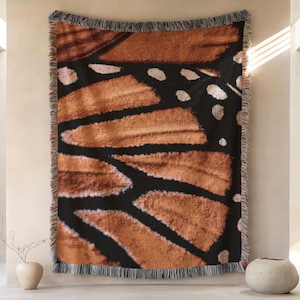 Op de afbeelding: Een geweven sprei met een gedetailleerd patroon van een monarchvlindervleugel. Het ontwerp toont bruine, zwarte en witte tinten, met een gefranjerde rand. De sprei wordt aan een muur getoond en biedt een op de natuur geïnspireerd decoratief element.