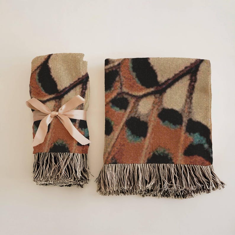 Op de afbeelding: Twee geweven sprei met een vlindervleugelpatroon. E&eacute;n deken is opgevouwen en vastgebonden met een roze lint. De andere deken is uitgevouwen. Beide dekens hebben een beige achtergrond met bruine, zwarte en turquoise accenten en franjes.