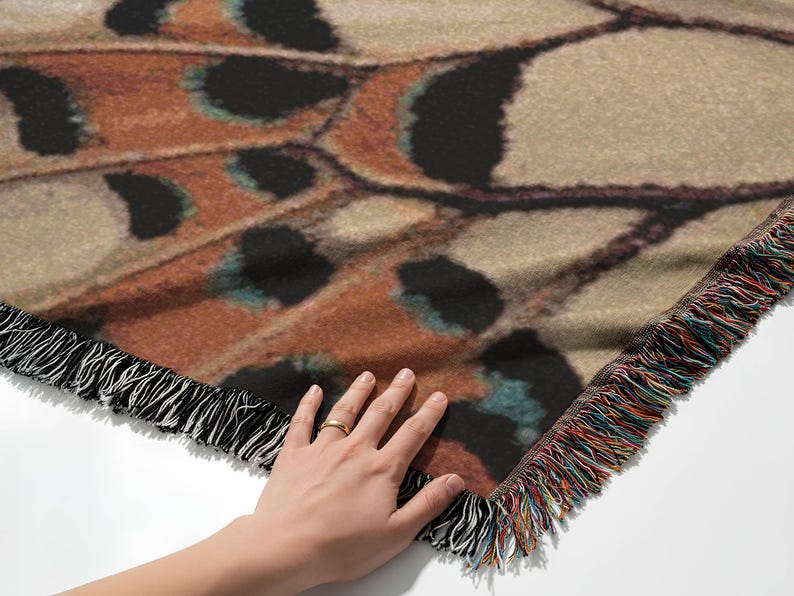 Op de afbeelding: Een sprei met een gedetailleerd vlindervleugelontwerp in bruin, beige en zwart. De deken heeft een gefranjerde rand met een mix van kleurrijke draden. De deken is gemaakt van een zacht materiaal.