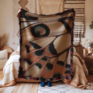 Op de afbeelding: Een geweven plaid met een gedetailleerde macro-print van een vlindervleugel in bruine, beige en zwarte tinten. De deken heeft een gestructureerd oppervlak en is afgewerkt met decoratieve franjes langs alle vier de randen in rood, zwart en wit.