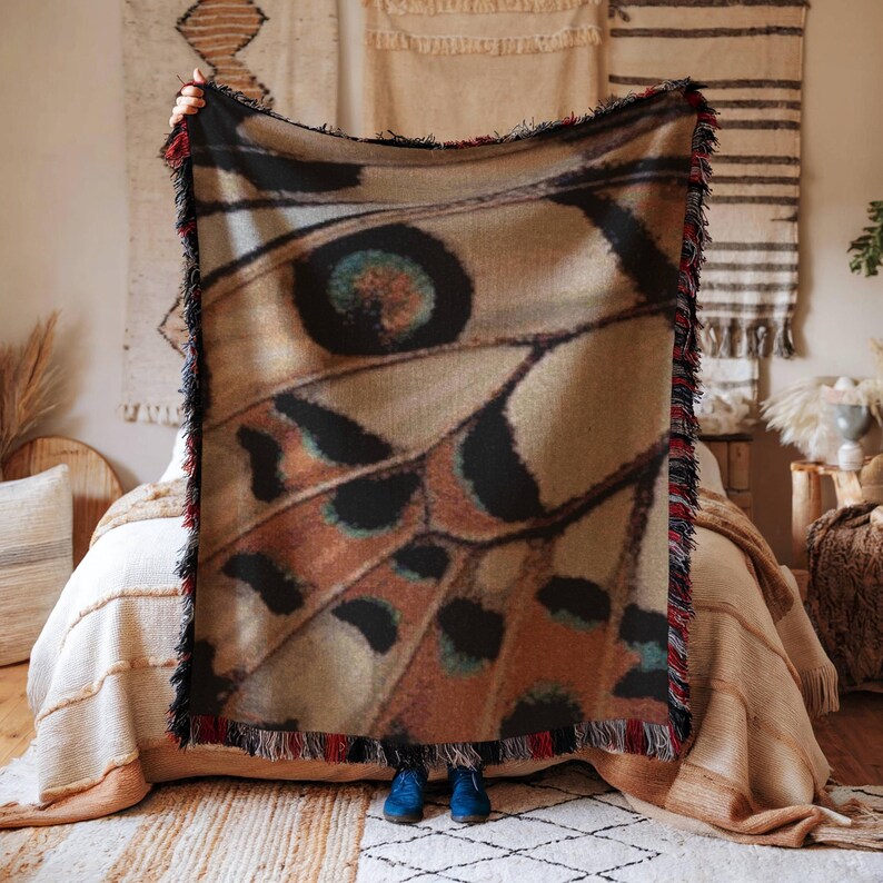 Op de afbeelding: Een geweven plaid met een gedetailleerde macro-print van een vlindervleugel in bruine, beige en zwarte tinten. De deken heeft een gestructureerd oppervlak en is afgewerkt met decoratieve franjes langs alle vier de randen in rood, zwart en wit.