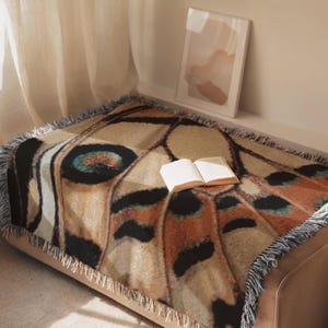 Op de afbeelding: Een geweven plaid met een gedetailleerd vlindervleugelpatroon in tinten bruin, beige, zwart en blauwgroen. De rechthoekige deken heeft een gefranjerde rand en ligt over een oppervlak met een open boek erop.