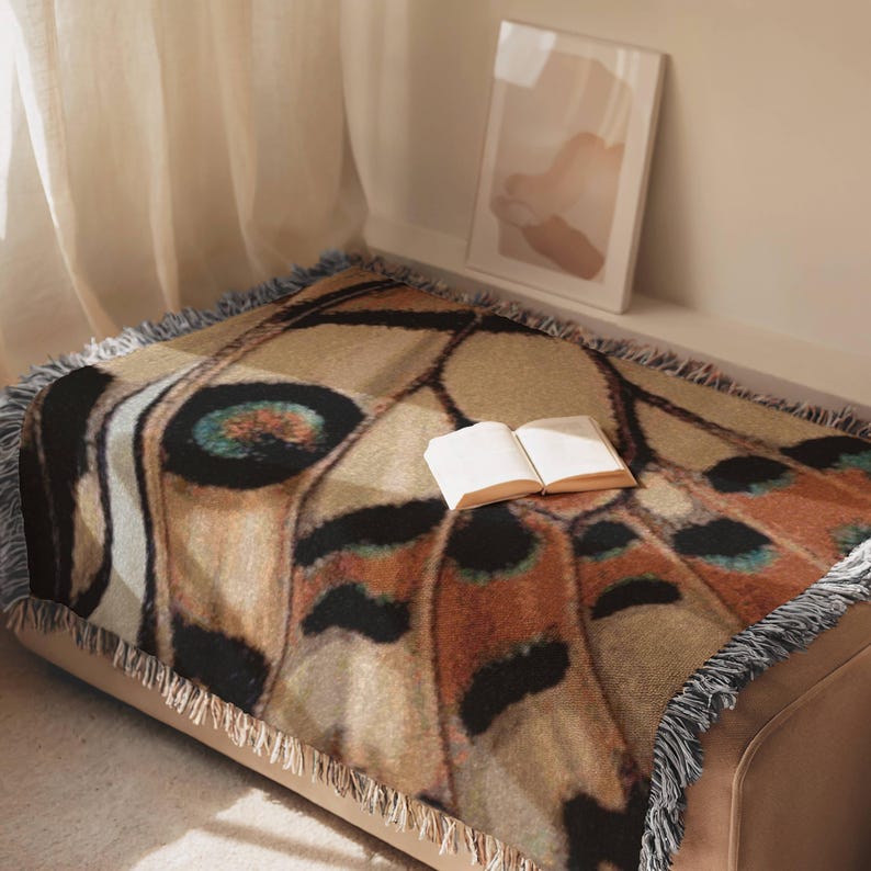 Op de afbeelding: Een geweven plaid met een gedetailleerd vlindervleugelpatroon in tinten bruin, beige, zwart en blauwgroen. De rechthoekige deken heeft een gefranjerde rand en ligt over een oppervlak met een open boek erop.