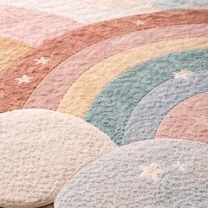 May include: A colorful, round rug with a rainbow design and cloud-shaped accents. The rug features pastel colors including pink, yellow, blue, and cream. White star details are scattered across the rug.