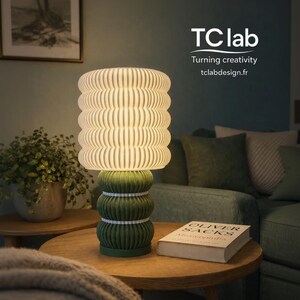 May include: A decorative table lamp with a ribbed, off-white lampshade and a green base. The lamp is on a wooden table with a book and a plant. The text "TC lab" and "Turning creativity" are visible.
