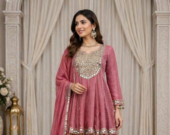 Könnte beinhalten: Staubrosa Salwar Kameez-Ensemble. Das Oberteil hat einen stark verzierten Ausschnitt und Manschetten mit aufwendiger Spiegelarbeit und goldenen Verzierungen. Die passende Hose ist im Dhoti-Stil, und ein Dupatta ist zur Seite drapiert. Festliche Kleidung.