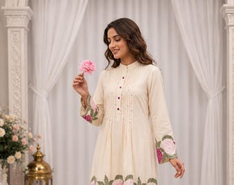 Könnte beinhalten: Ein cremefarbenes Tunika- und Hosenset mit Stehkragen, vertikalen Falten und dunkelrosa Knöpfen. Das Kleidungsstück ist mit gestickten rosa und hellrosa Blumenmotiven sowie grünen Blättern an den Ärmeln und am Saum verziert.