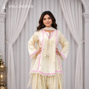 K&ouml;nnte beinhalten: Ein cremefarbenes Salwar-Kameez-Set bestehend aus einer Tunika mit vertikalen Streifen, rosa Spitzenbesatz und einem Schn&uuml;rhalsausschnitt mit Quasten. Die passende weite Hose und der transparente Dupatta sind mit einem zarten Blumenmuster verziert.