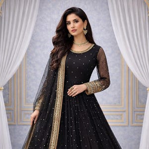 Peut inclure: Une robe de soir&eacute;e noire orn&eacute;e de d&eacute;tails dor&eacute;s. La robe pr&eacute;sente une longue jupe et des manches en tissu transparent. Des bordures dor&eacute;es ornent l'encolure, les poignets et l'ourlet. La mod&egrave;le porte des bijoux dor&eacute;s.